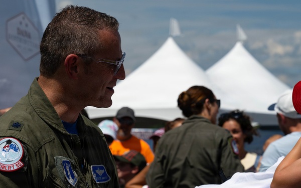 A military aviator in an olive green flight suit is signing autographs at what appears to be the New York Air Show. He is signing a white t-shirt that reads 'Air Dot Show New York' being held by a child wearing a red baseball cap. The event is taking place outdoors with white event tents visible in the background and a crowd of attendees.