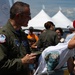 A military aviator in an olive green flight suit is signing autographs at what appears to be the New York Air Show. He is signing a white t-shirt that reads 'Air Dot Show New York' being held by a child wearing a red baseball cap. The event is taking place outdoors with white event tents visible in the background and a crowd of attendees.
