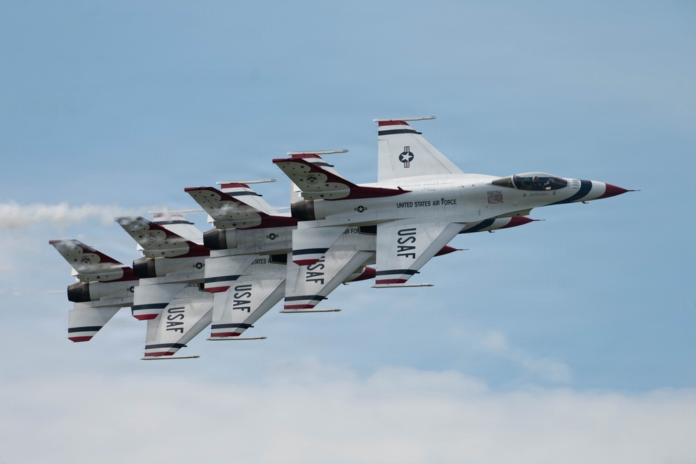 The image shows a formation of United States Air Force Thunderbirds flying in close formation against a partly cloudy blue sky. The aircraft are F-16 Fighting Falcons painted in the iconic Thunderbirds livery of white, red, and blue. At least three aircraft are visible flying in a tight echelon formation, with light contrail/exhaust visible from the engines.