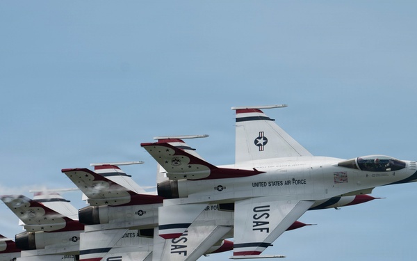 The image shows a formation of United States Air Force Thunderbirds flying in close formation against a partly cloudy blue sky. The aircraft are F-16 Fighting Falcons painted in the iconic Thunderbirds livery of white, red, and blue. At least three aircraft are visible flying in a tight echelon formation, with light contrail/exhaust visible from the engines.