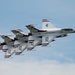 The image shows a formation of United States Air Force Thunderbirds flying in close formation against a partly cloudy blue sky. The aircraft are F-16 Fighting Falcons painted in the iconic Thunderbirds livery of white, red, and blue. At least three aircraft are visible flying in a tight echelon formation, with light contrail/exhaust visible from the engines.