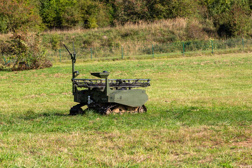 A small military-green unmanned ground vehicle (UGV) is positioned in an open grassy field. The tracked robotic platform appears to be equipped with sensors, antennas, and a payload rack on top. The setting suggests a field demonstration or testing environment.