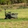 A small military-green unmanned ground vehicle (UGV) is positioned in an open grassy field. The tracked robotic platform appears to be equipped with sensors, antennas, and a payload rack on top. The setting suggests a field demonstration or testing environment.