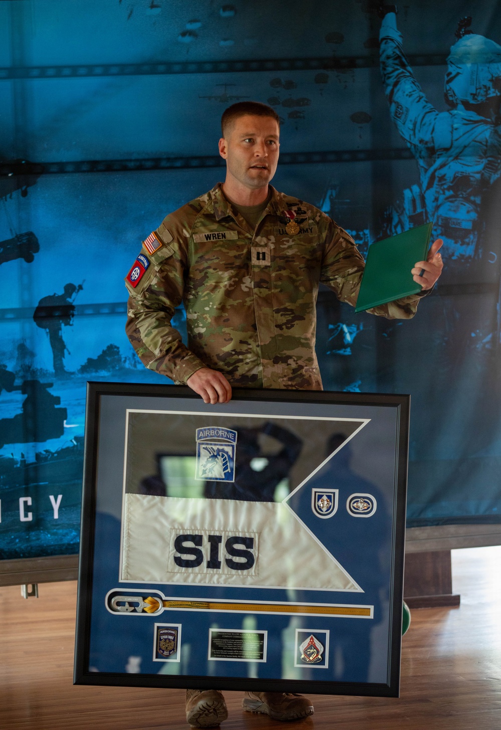 A U.S. Army Captain named Wren, wearing OCP (Operational Camouflage Pattern) uniform with 82nd Airborne Division patch and a medal on his chest, stands holding a framed shadow box display. The shadow box contains a military guidon flag marked 'SIS', an Airborne patch with a horse insignia, various unit patches, and what appears to be a saber or sword. This appears to be a military awards or farewell ceremony.