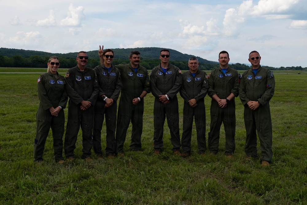 A group of eight military personnel in olive/dark green flight suits are posing for a group photo on a grassy airfield. They are wearing matching uniforms with patches and blue insignia, suggesting they belong to the same unit or squadron. The setting appears to be an outdoor airfield with rolling hills and partly cloudy skies in the background.
