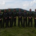 A group of eight military personnel in olive/dark green flight suits are posing for a group photo on a grassy airfield. They are wearing matching uniforms with patches and blue insignia, suggesting they belong to the same unit or squadron. The setting appears to be an outdoor airfield with rolling hills and partly cloudy skies in the background.