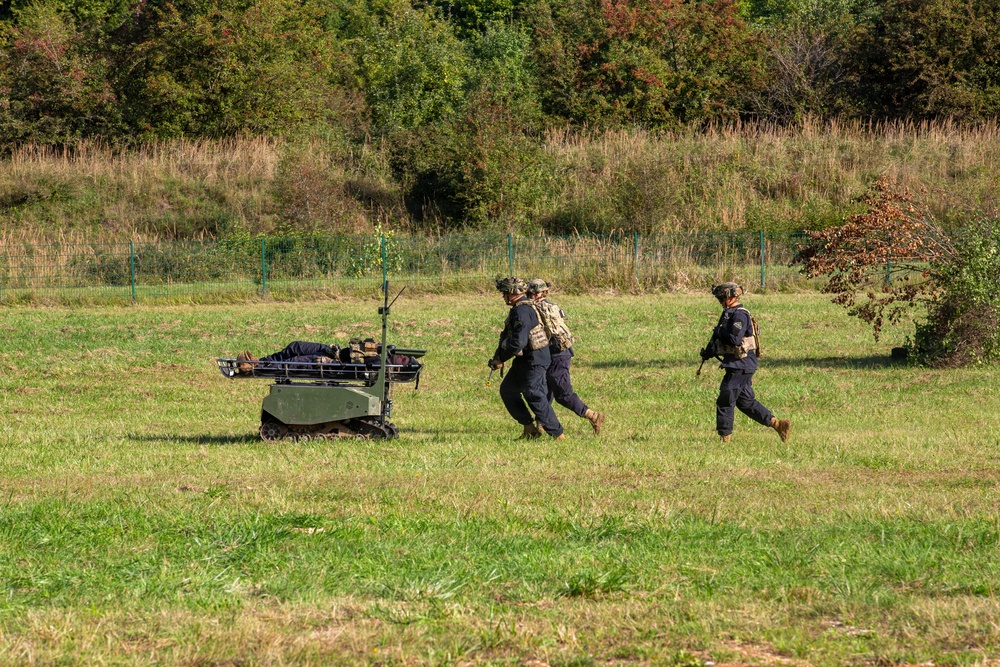 The image shows a military or tactical exercise in an open grassy field. Two personnel in dark uniforms and helmets are moving across the field alongside a small unmanned ground vehicle (UGV) that appears to be carrying a simulated casualty on a stretcher platform. The scene suggests a medical evacuation or casualty extraction drill using robotic assistance.