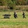 The image shows a military or tactical exercise in an open grassy field. Two personnel in dark uniforms and helmets are moving across the field alongside a small unmanned ground vehicle (UGV) that appears to be carrying a simulated casualty on a stretcher platform. The scene suggests a medical evacuation or casualty extraction drill using robotic assistance.