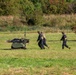 The image shows a military or tactical exercise in an open grassy field. Two personnel in dark uniforms and helmets are moving across the field alongside a small unmanned ground vehicle (UGV) that appears to be carrying a simulated casualty on a stretcher platform. The scene suggests a medical evacuation or casualty extraction drill using robotic assistance.