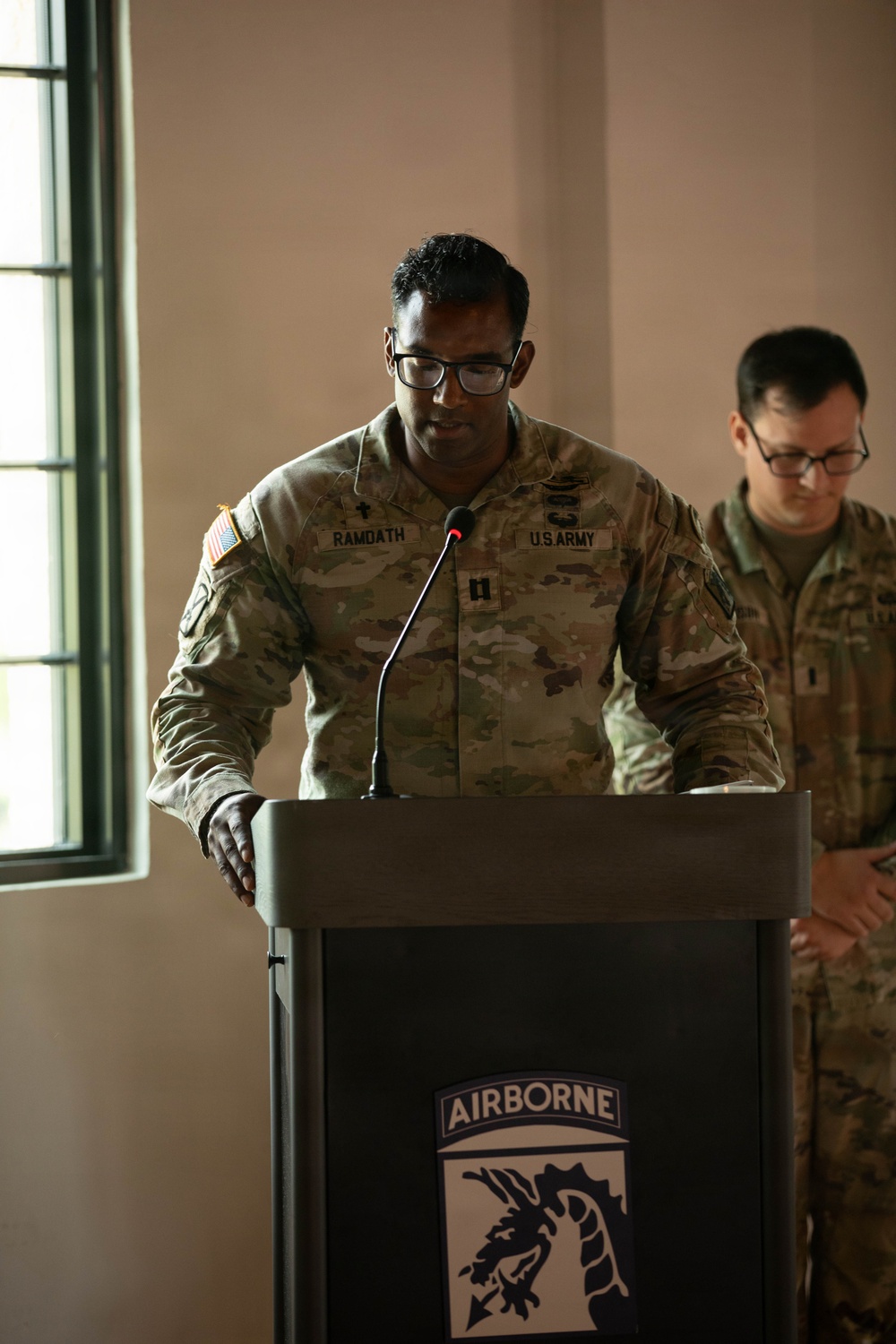 A U.S. Army soldier named Ramdath, wearing the rank of Captain and a Chaplain's cross insignia, stands at a podium bearing the 18th Airborne Corps insignia. He appears to be delivering a prayer or address at a formal military ceremony. A second soldier stands behind him with hands clasped, head bowed.