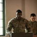 A U.S. Army soldier named Ramdath, wearing the rank of Captain and a Chaplain's cross insignia, stands at a podium bearing the 18th Airborne Corps insignia. He appears to be delivering a prayer or address at a formal military ceremony. A second soldier stands behind him with hands clasped, head bowed.