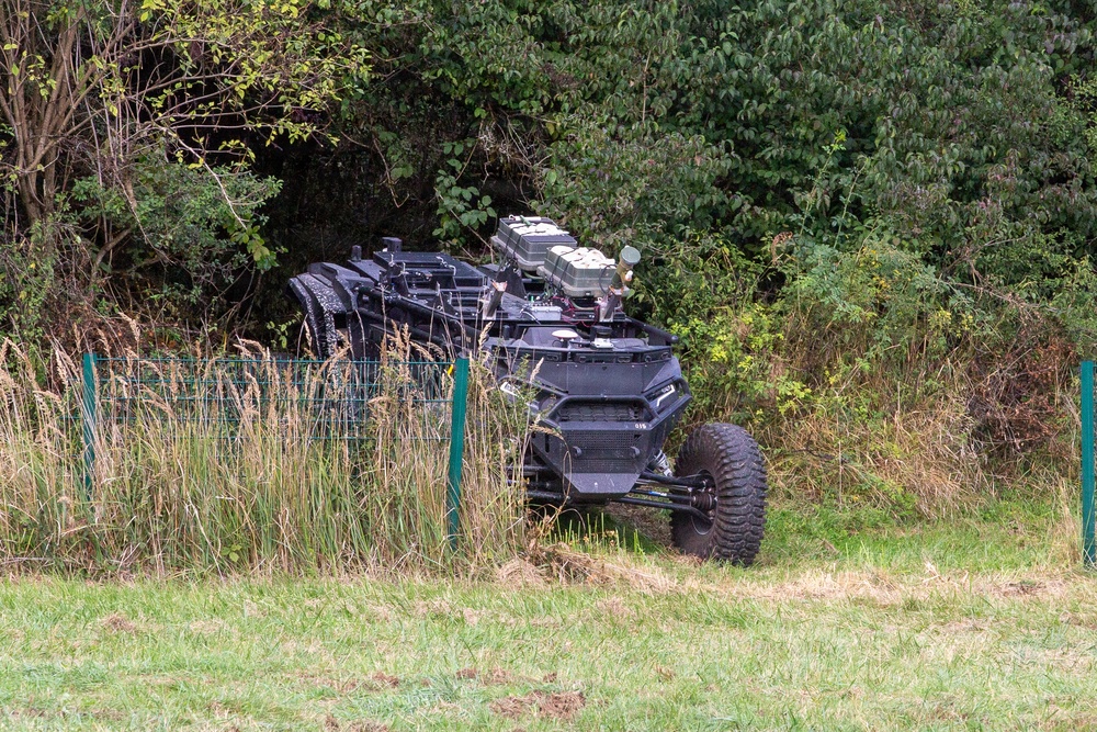 A dark-colored military or tactical off-road vehicle is partially concealed among tall grass, shrubs, and trees near a green wire fence. The vehicle appears to be a wheeled unmanned ground vehicle (UGV) or tactical utility vehicle equipped with electronic equipment or sensor modules on top. The setting appears to be a rural or training area with mixed vegetation.