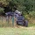 A dark-colored military or tactical off-road vehicle is partially concealed among tall grass, shrubs, and trees near a green wire fence. The vehicle appears to be a wheeled unmanned ground vehicle (UGV) or tactical utility vehicle equipped with electronic equipment or sensor modules on top. The setting appears to be a rural or training area with mixed vegetation.
