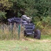 A dark-colored military or tactical off-road vehicle is partially concealed among tall grass, shrubs, and trees near a green wire fence. The vehicle appears to be a wheeled unmanned ground vehicle (UGV) or tactical utility vehicle equipped with electronic equipment or sensor modules on top. The setting appears to be a rural or training area with mixed vegetation.