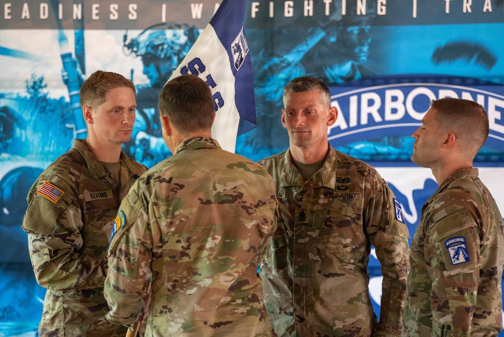 Four U.S. Army soldiers in OCP (Operational Camouflage Pattern) uniforms are gathered during what appears to be a military ceremony, likely a change of command or unit activation event. A blue and white unit guidon flag is visible in the background along with a large banner displaying 'AIRBORNE' and words including 'READINESS,' 'WARFIGHTING,' and 'TRAINING.' The soldier facing away appears to be presenting or passing the guidon flag.