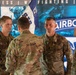 Four U.S. Army soldiers in OCP (Operational Camouflage Pattern) uniforms are gathered during what appears to be a military ceremony, likely a change of command or unit activation event. A blue and white unit guidon flag is visible in the background along with a large banner displaying 'AIRBORNE' and words including 'READINESS,' 'WARFIGHTING,' and 'TRAINING.' The soldier facing away appears to be presenting or passing the guidon flag.