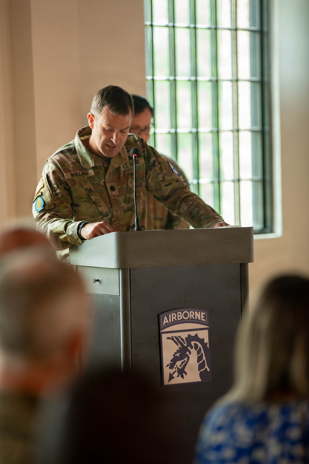 A U.S. Army soldier with the name tape 'MULLANY' is speaking at a podium bearing the 18th Airborne Corps insignia. The soldier is wearing an OCP (Operational Camouflage Pattern) uniform and appears to be delivering remarks at an official military ceremony or event. An audience is visible in the foreground, slightly out of focus.