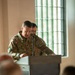 A U.S. Army soldier with the name tape 'MULLANY' is speaking at a podium bearing the 18th Airborne Corps insignia. The soldier is wearing an OCP (Operational Camouflage Pattern) uniform and appears to be delivering remarks at an official military ceremony or event. An audience is visible in the foreground, slightly out of focus.