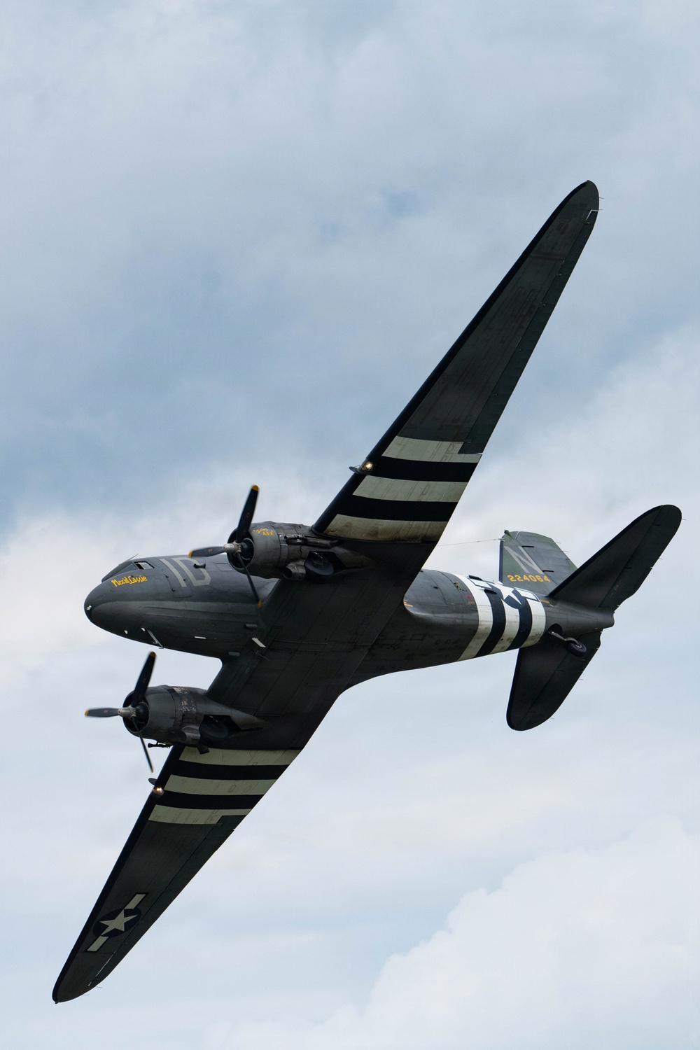 A World War II-era Douglas C-47 Skytrain military transport aircraft is photographed in flight from below against a partly cloudy sky. The aircraft displays D-Day invasion stripes (alternating black and white bands) on its wings and fuselage, along with USAAF markings. The nose art reads 'Placid Lassie' and the tail number '224064' is visible, with the fuselage code '1C'.