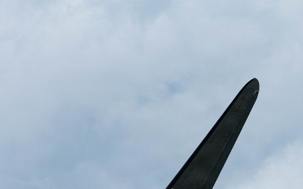 A World War II-era Douglas C-47 Skytrain military transport aircraft is photographed in flight from below against a partly cloudy sky. The aircraft displays D-Day invasion stripes (alternating black and white bands) on its wings and fuselage, along with USAAF markings. The nose art reads 'Placid Lassie' and the tail number '224064' is visible, with the fuselage code '1C'.