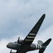 A World War II-era Douglas C-47 Skytrain military transport aircraft is photographed in flight from below against a partly cloudy sky. The aircraft displays D-Day invasion stripes (alternating black and white bands) on its wings and fuselage, along with USAAF markings. The nose art reads 'Placid Lassie' and the tail number '224064' is visible, with the fuselage code '1C'.