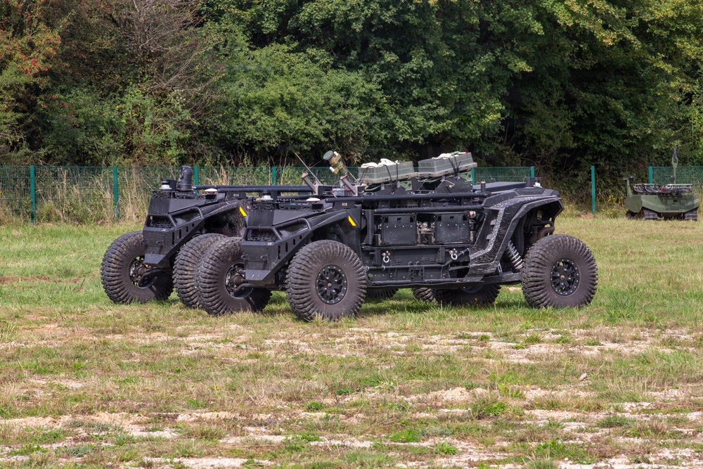 Two black unmanned ground vehicles (UGVs) are parked side by side on a grassy field, appearing to be military robotic platforms. Both vehicles feature large off-road tires, open chassis frames, and sensor/equipment mounts on top. A third vehicle is partially visible in the background to the right.
