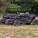 Two black unmanned ground vehicles (UGVs) are parked side by side on a grassy field, appearing to be military robotic platforms. Both vehicles feature large off-road tires, open chassis frames, and sensor/equipment mounts on top. A third vehicle is partially visible in the background to the right.