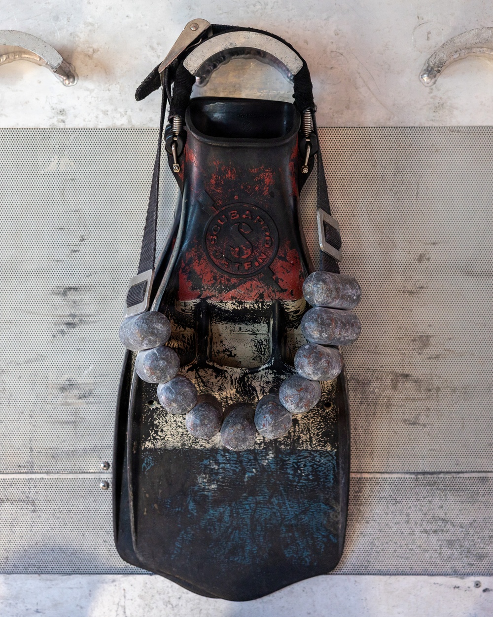 The image shows a heavily worn and weathered scuba diving fin, branded 'Scubapro Delfino,' mounted or laid flat on a perforated metal surface. The fin has significant paint wear revealing layers of red, black, and gold coloring, and is adorned with a string of rounded gray beads or weights arranged in a U-shape across the blade. The overall condition suggests extensive use and age.