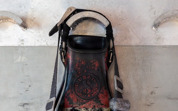 The image shows a heavily worn and weathered scuba diving fin, branded 'Scubapro Delfino,' mounted or laid flat on a perforated metal surface. The fin has significant paint wear revealing layers of red, black, and gold coloring, and is adorned with a string of rounded gray beads or weights arranged in a U-shape across the blade. The overall condition suggests extensive use and age.