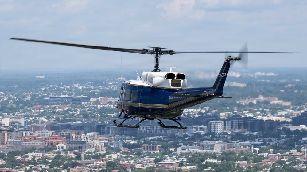 A blue and white military/government helicopter is captured in flight over a dense urban landscape. The helicopter displays the tail number '96667' and bears markings consistent with United States government or military use. The city below features a mix of residential and commercial buildings with abundant tree cover, consistent with the Washington D.C. metropolitan area.
