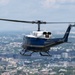 A blue and white military/government helicopter is captured in flight over a dense urban landscape. The helicopter displays the tail number '96667' and bears markings consistent with United States government or military use. The city below features a mix of residential and commercial buildings with abundant tree cover, consistent with the Washington D.C. metropolitan area.