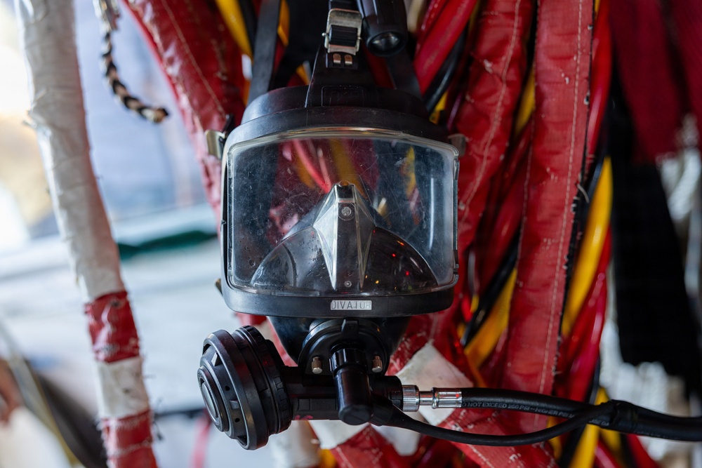 The image shows a full-face diving mask, branded 'DIVATOR', hanging against a background of red and yellow equipment straps or hoses. The mask features a large rectangular visor, a demand valve/regulator assembly at the bottom, and connected hoses or cables. The equipment appears to be professional-grade commercial or rescue diving gear stored or mounted in a working environment.