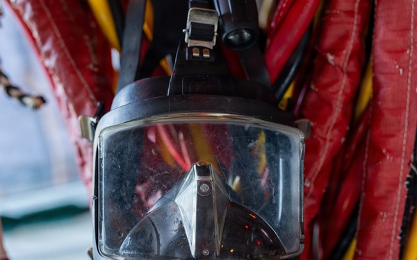 The image shows a full-face diving mask, branded 'DIVATOR', hanging against a background of red and yellow equipment straps or hoses. The mask features a large rectangular visor, a demand valve/regulator assembly at the bottom, and connected hoses or cables. The equipment appears to be professional-grade commercial or rescue diving gear stored or mounted in a working environment.