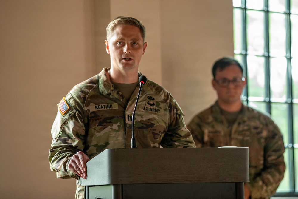 A U.S. Army soldier identified by name tape as 'KEATING' is speaking at a podium with a microphone, wearing Operational Camouflage Pattern (OCP) uniform with a U.S. flag patch on the left shoulder. A second soldier in the background, partially visible and out of focus, appears to be observing the proceedings. The setting appears to be an indoor military ceremony or briefing.