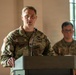A U.S. Army soldier identified by name tape as 'KEATING' is speaking at a podium with a microphone, wearing Operational Camouflage Pattern (OCP) uniform with a U.S. flag patch on the left shoulder. A second soldier in the background, partially visible and out of focus, appears to be observing the proceedings. The setting appears to be an indoor military ceremony or briefing.