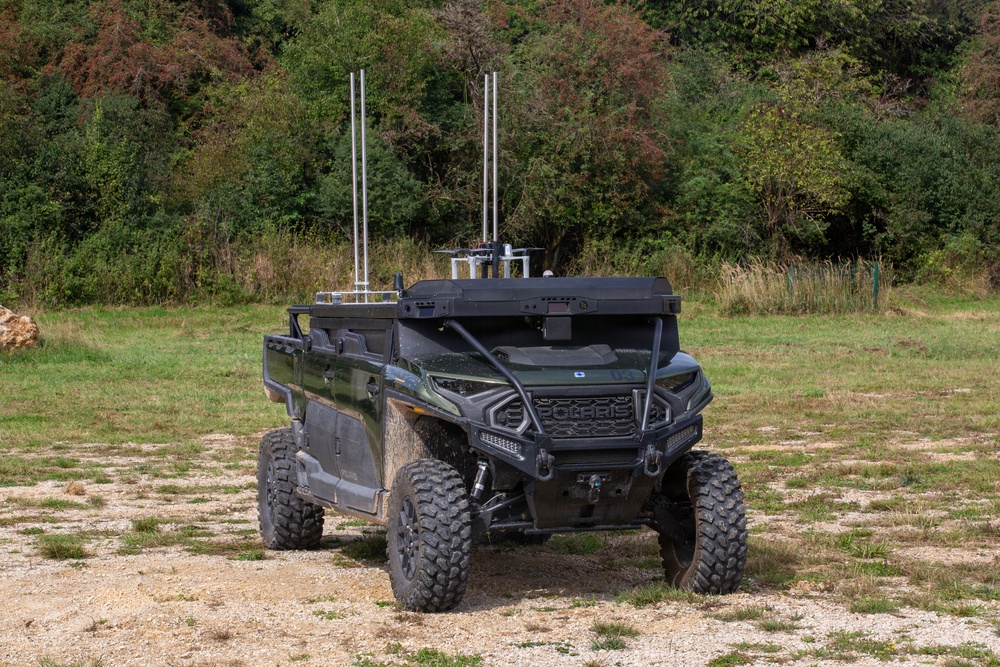 A military-configured Polaris utility terrain vehicle (UTV) is parked on a gravel and grass clearing with dense tree line in the background. The vehicle has been modified with a roof-mounted equipment platform featuring two vertical antennas and additional electronic systems. The vehicle appears to be an unmanned ground vehicle (UGV) or remotely operated platform based on the absence of visible occupants and the sensor/antenna array.
