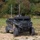 A military-configured Polaris utility terrain vehicle (UTV) is parked on a gravel and grass clearing with dense tree line in the background. The vehicle has been modified with a roof-mounted equipment platform featuring two vertical antennas and additional electronic systems. The vehicle appears to be an unmanned ground vehicle (UGV) or remotely operated platform based on the absence of visible occupants and the sensor/antenna array.