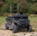 A military-configured Polaris utility terrain vehicle (UTV) is parked on a gravel and grass clearing with dense tree line in the background. The vehicle has been modified with a roof-mounted equipment platform featuring two vertical antennas and additional electronic systems. The vehicle appears to be an unmanned ground vehicle (UGV) or remotely operated platform based on the absence of visible occupants and the sensor/antenna array.