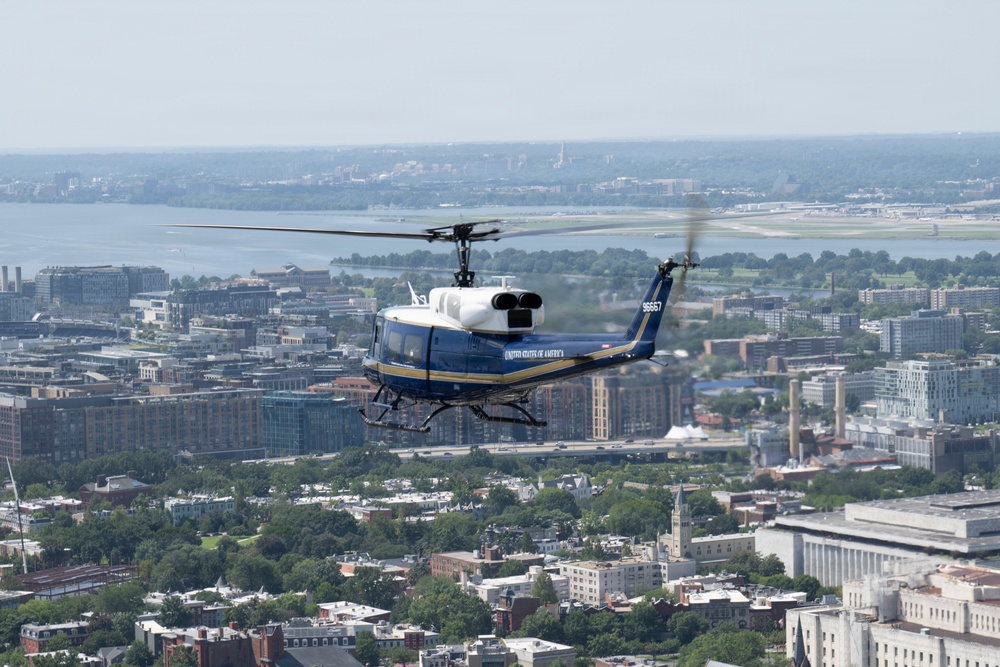 A United States of America military helicopter, tail number 96557, flies over what appears to be Washington D.C., with the Potomac River and Reagan National Airport visible in the background. The helicopter is a blue and white UH-1 'Huey' variant in executive transport livery, consistent with those used by the U.S. Army for VIP transport. The urban landscape below includes dense city blocks, tree-lined streets, and federal-style buildings.