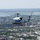A United States of America military helicopter, tail number 96557, flies over what appears to be Washington D.C., with the Potomac River and Reagan National Airport visible in the background. The helicopter is a blue and white UH-1 'Huey' variant in executive transport livery, consistent with those used by the U.S. Army for VIP transport. The urban landscape below includes dense city blocks, tree-lined streets, and federal-style buildings.