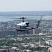 A United States of America military helicopter, tail number 96557, flies over what appears to be Washington D.C., with the Potomac River and Reagan National Airport visible in the background. The helicopter is a blue and white UH-1 'Huey' variant in executive transport livery, consistent with those used by the U.S. Army for VIP transport. The urban landscape below includes dense city blocks, tree-lined streets, and federal-style buildings.