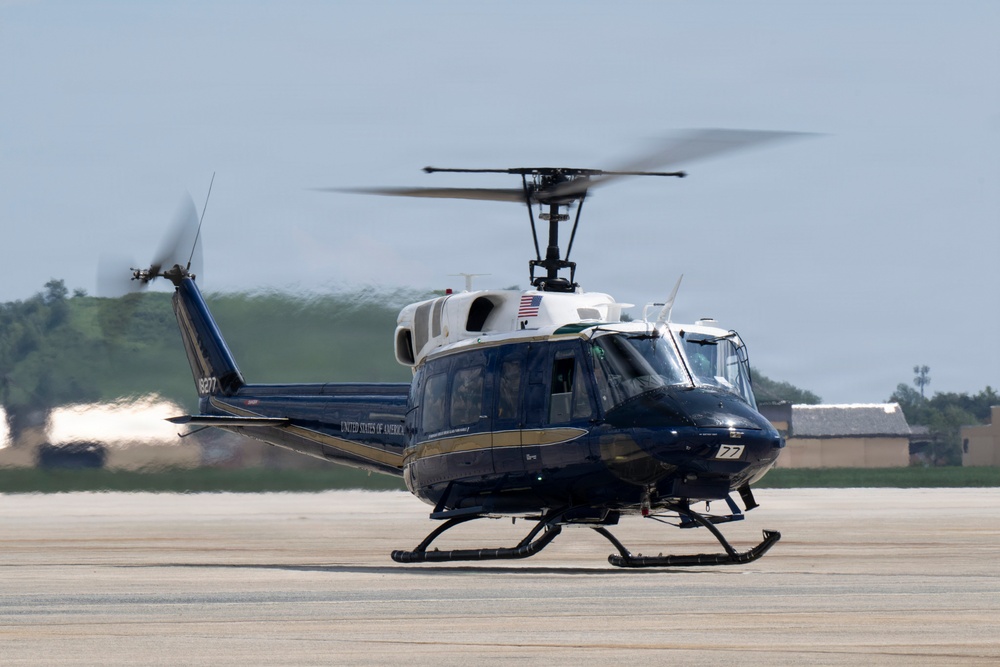 A dark navy blue and white United States of America government helicopter is captured on a tarmac, appearing to be in the process of landing or taking off. The aircraft bears the inscription 'United States of America' along its fuselage and displays an American flag near the rotor assembly. The rotor blades are visibly in motion, shown by motion blur, indicating active operation.