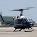 A dark navy blue and white United States of America government helicopter is captured on a tarmac, appearing to be in the process of landing or taking off. The aircraft bears the inscription 'United States of America' along its fuselage and displays an American flag near the rotor assembly. The rotor blades are visibly in motion, shown by motion blur, indicating active operation.