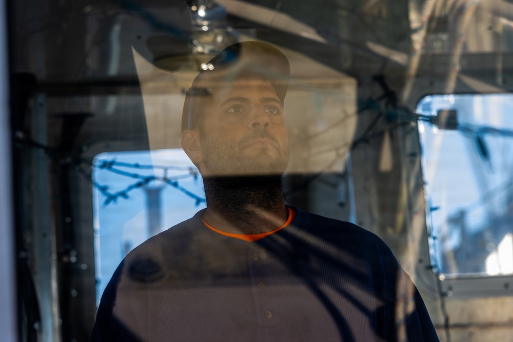 A man is photographed through a glass or transparent surface, creating reflections and layered visual effects. He appears to be inside an enclosed space, wearing a dark shirt with an orange collar. The image has a contemplative, somewhat somber mood with industrial or institutional elements visible in the background.