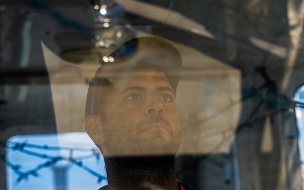 A man is photographed through a glass or transparent surface, creating reflections and layered visual effects. He appears to be inside an enclosed space, wearing a dark shirt with an orange collar. The image has a contemplative, somewhat somber mood with industrial or institutional elements visible in the background.