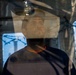 A man is photographed through a glass or transparent surface, creating reflections and layered visual effects. He appears to be inside an enclosed space, wearing a dark shirt with an orange collar. The image has a contemplative, somewhat somber mood with industrial or institutional elements visible in the background.