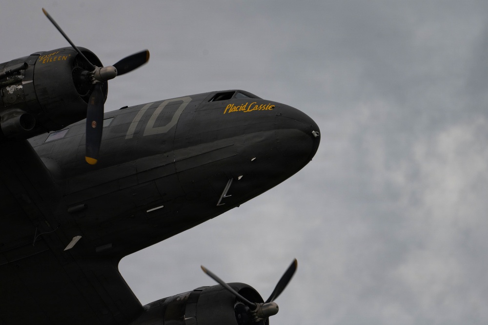 A dark olive/black painted WWII-era C-47 Skytrain transport aircraft photographed from a low angle against an overcast sky. The aircraft bears the nose art name 'Placid Cassie' in yellow lettering and the number '10' on its fuselage. A second aircraft, partially visible in the lower frame, bears the name 'Lady Eileen' near its engine cowling.