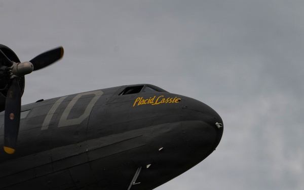 A dark olive/black painted WWII-era C-47 Skytrain transport aircraft photographed from a low angle against an overcast sky. The aircraft bears the nose art name 'Placid Cassie' in yellow lettering and the number '10' on its fuselage. A second aircraft, partially visible in the lower frame, bears the name 'Lady Eileen' near its engine cowling.