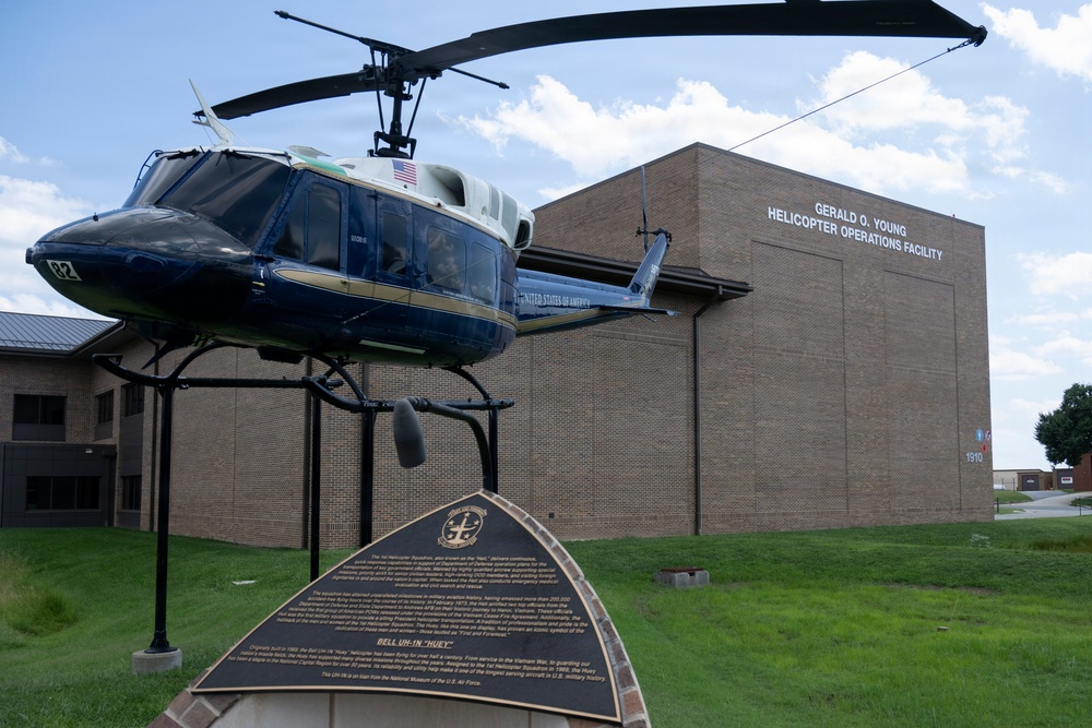 A Bell UH-1 'Huey' helicopter is displayed on a static mount outside the Gerald O. Young Helicopter Operations Facility, a brick military building. The helicopter bears 'United States of America' markings and an American flag, and is accompanied by an informational placard at its base. The scene appears to be a military base or aviation museum outdoor exhibit.