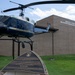 A Bell UH-1 'Huey' helicopter is displayed on a static mount outside the Gerald O. Young Helicopter Operations Facility, a brick military building. The helicopter bears 'United States of America' markings and an American flag, and is accompanied by an informational placard at its base. The scene appears to be a military base or aviation museum outdoor exhibit.
